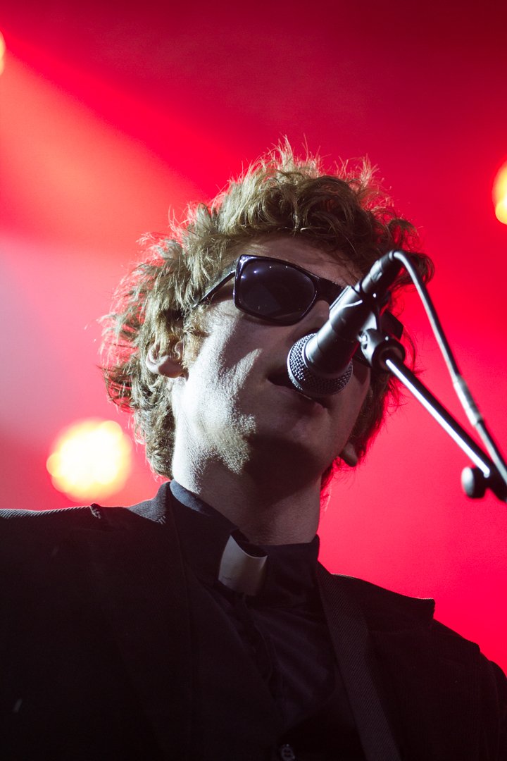 Ein Musiker mit Sonnenbrille und schwarzem Hemd mit weißem Kragen singt und spielt Gitarre vor roten Bühnenlichtern.