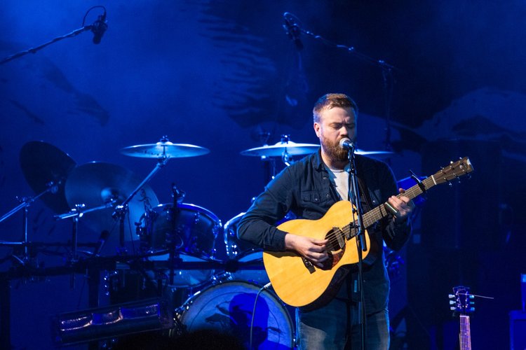 Ein bärtiger Musiker singt und spielt Akustikgitarre unter blauem Licht, vor einem Schlagzeug und einer Fischskelett-Dekoration.