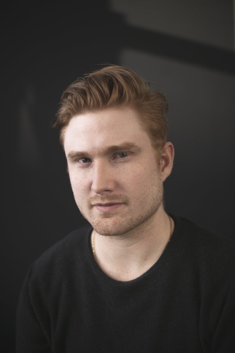 Ein junger Mann mit rötlichen Haaren, leichtem Bart und schwarzem Hemd vor dunklem Hintergrund.