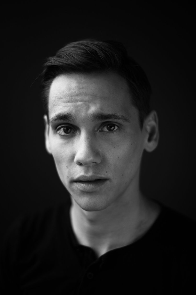 Schwarzweißporträt eines jungen Mannes mit kurzen Haaren, der vor dunklem Hintergrund in die Kamera blickt.
