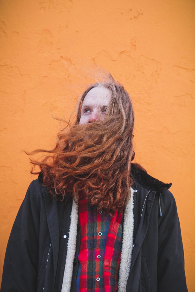 Eine junge Frau mit langen roten Haaren und kariertem Hemd vor einer orangefarbenen Wand.