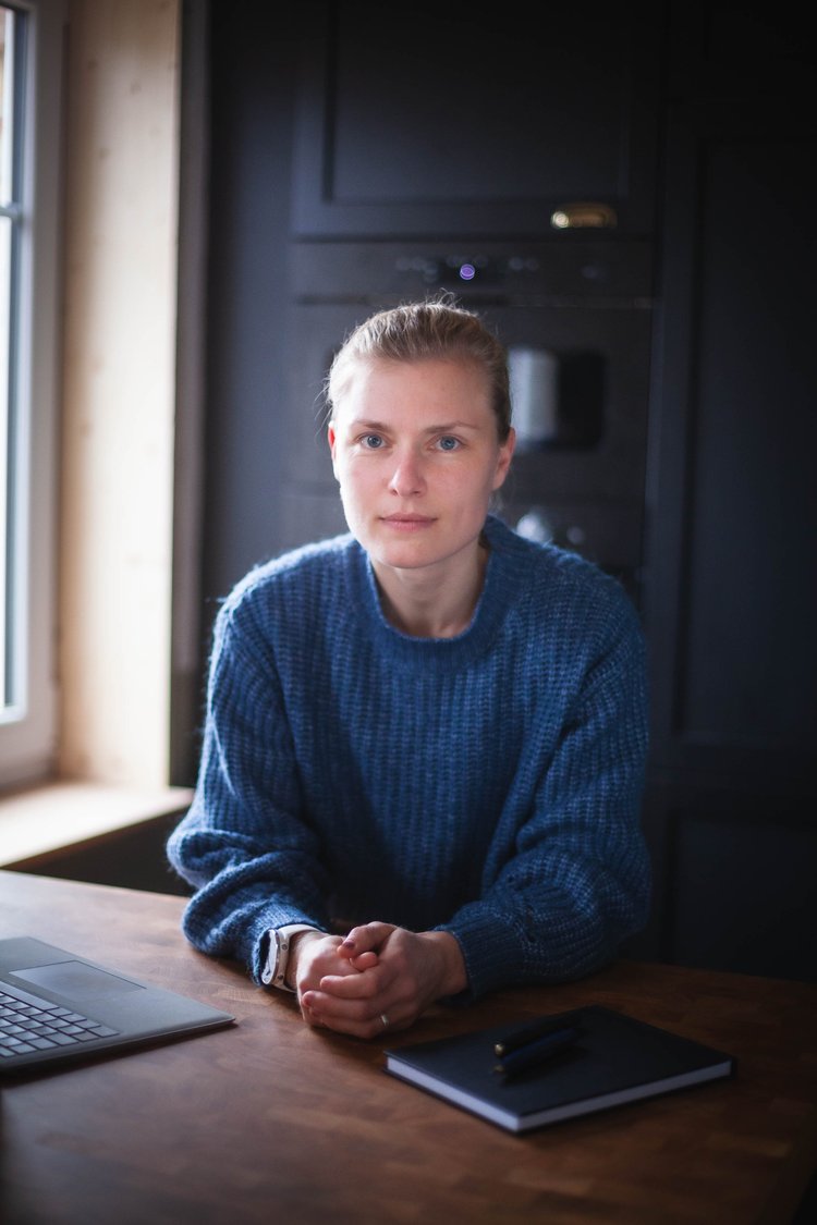 Eine Frau mit hellen Haaren und blauem Pullover sitzt an einem Holztisch vor dunklen Schränken und einem Fenster.