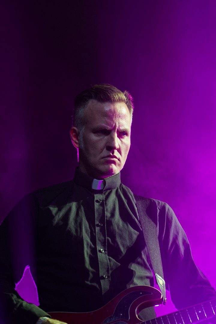 Ein als Priester gekleideter Mann spielt E-Gitarre unter violettem Bühnenlicht.