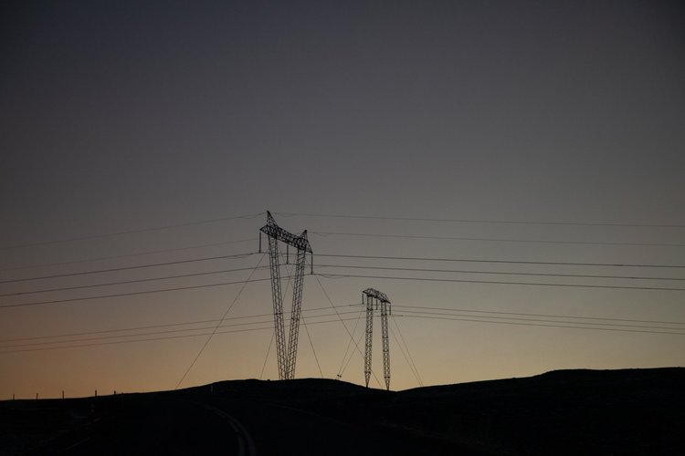 Silhouetten von Strommasten vor einem dämmrigen Himmel und sanften Hügeln.