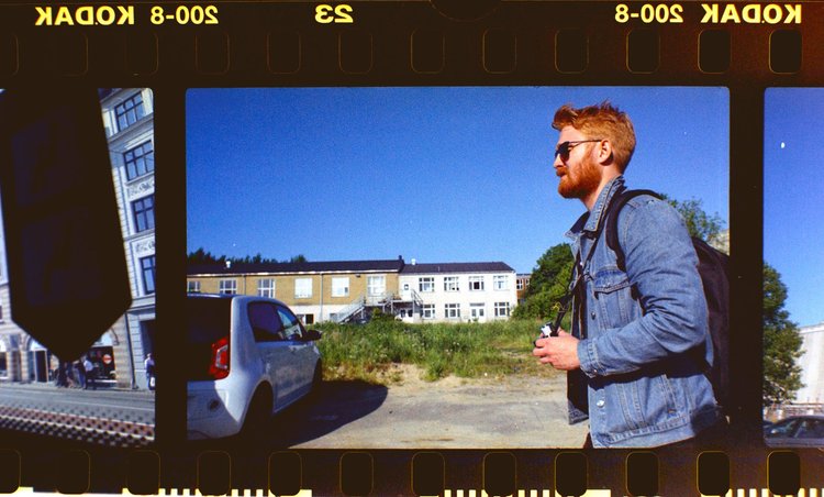 Ein junger Mann mit roten Haaren, Bart, Sonnenbrille, Jeansjacke und Rucksack unterwegs an einem sonnigen Tag.