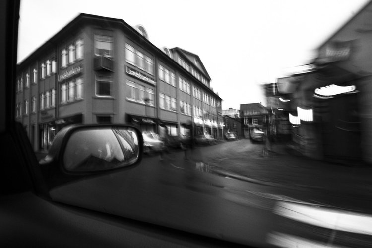 Schwarzweiße Straßenszene aus einem Fahrzeug heraus, mit unscharfen Gebäuden, Autos und einer Spiegelung im Seitenspiegel.