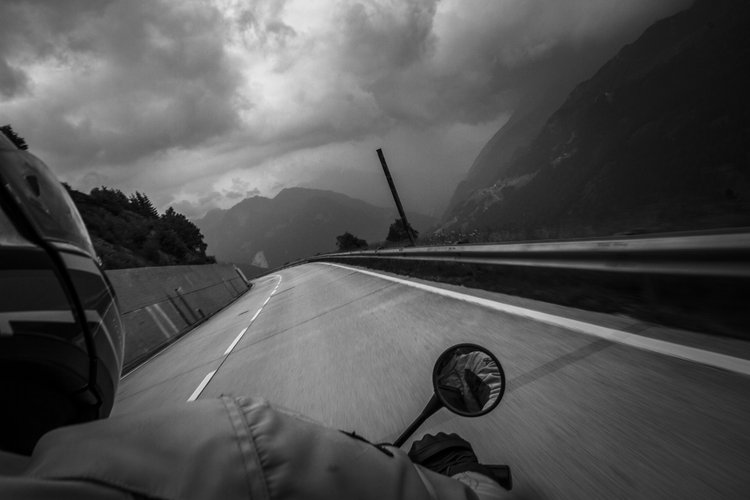 Schwarzweißaufnahme eines Motorradfahrers auf einer kurvigen Bergstraße unter dunklen Wolken.