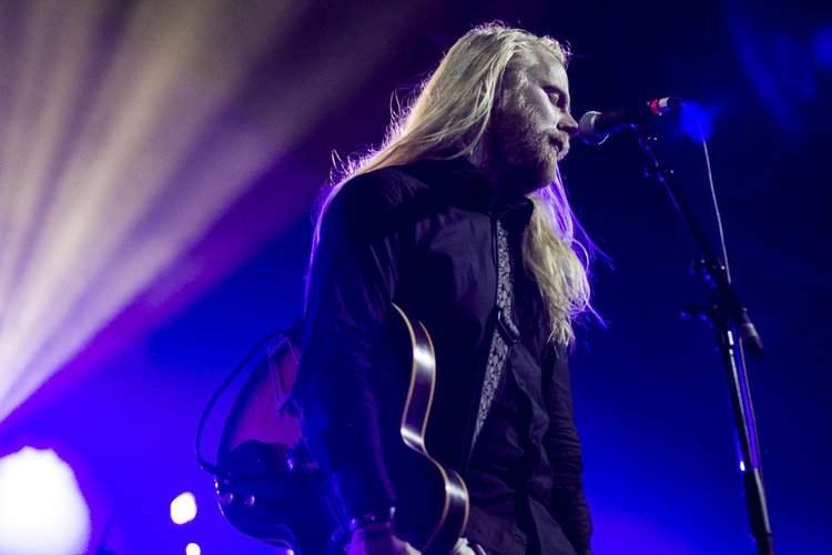 Ein Musiker mit langen blonden Haaren und Bart an Mikrofon und Gitarre unter violettem und blauem Bühnenlicht.