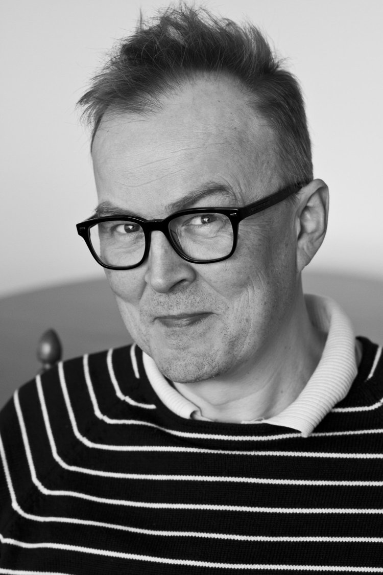 Schwarzweißporträt eines leicht lächelnden Mannes mit Brille und gestreiftem Pullover.