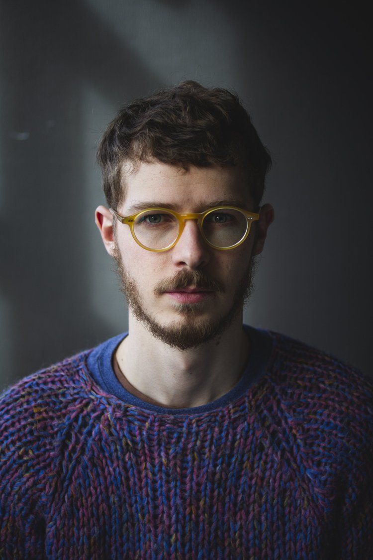 Ein junger Mann mit braunen Haaren, Bart, gelber Brille und buntem Strickpullover vor dunklem Hintergrund.
