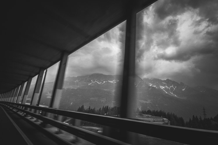 Schwarzweiße Berglandschaft durch ein Busfenster, mit einer Absperrung und Trägern im Vordergrund.