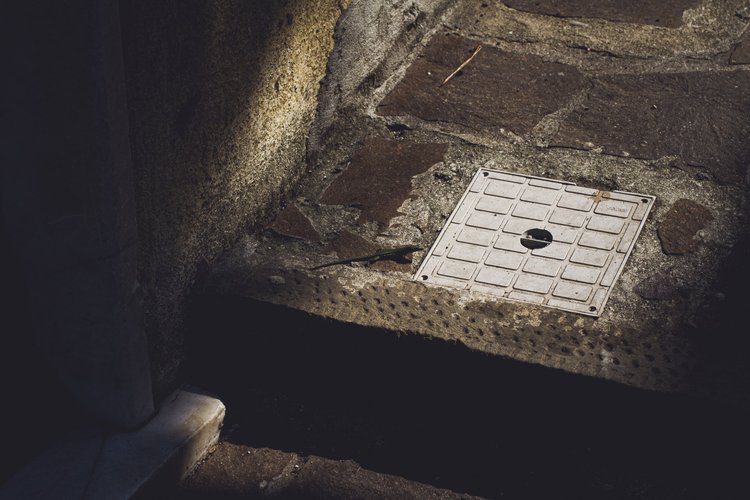Ein Abflussgitter in einem gepflasterten Gehweg, umgeben von Steinen und Beton.