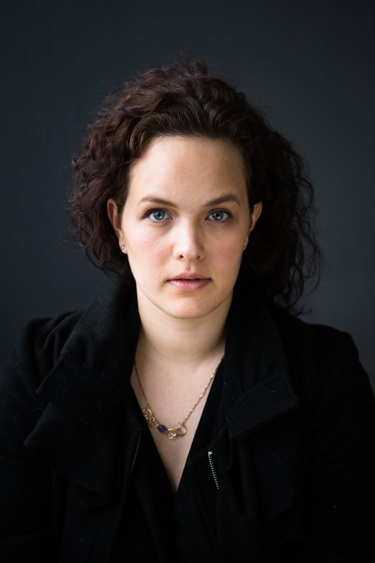 Porträt einer Frau mit braunen Locken, blauen Augen, schwarzer Jacke und goldener Halskette vor dunkelgrauem Hintergrund.