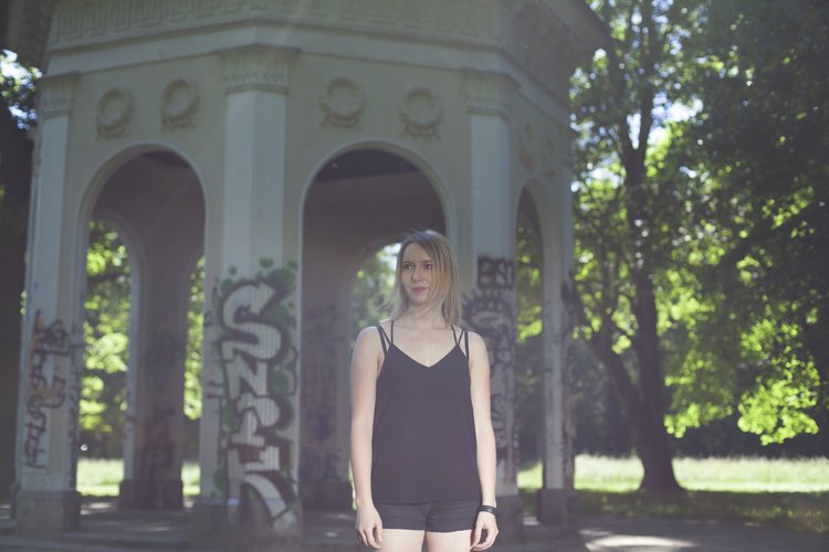 Eine junge Frau mit schwarzem Oberteil und Shorts steht in einem grünen Park vor einem weißen, mit Graffiti bedeckten Pavillon.