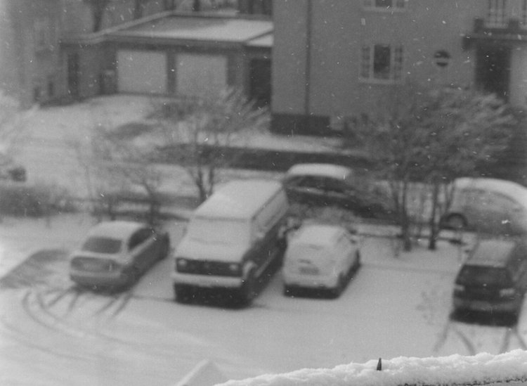 Ein verschneiter Parkplatz mit Autos, Bäumen und Wohnhäusern.