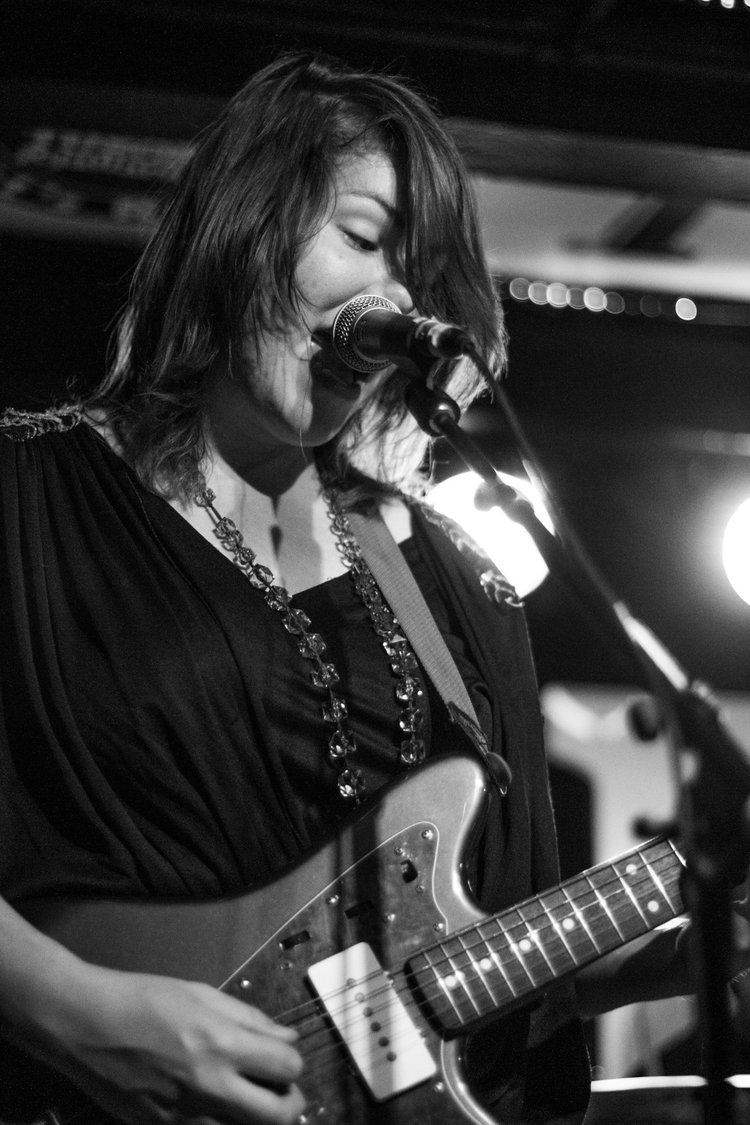Eine Frau singt am Mikrofon und spielt E-Gitarre. Schwarzweißaufnahme eines Liveauftritts.