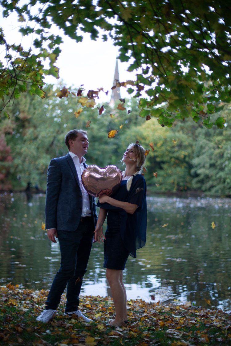 Ein Paar am Wasser mit einem herzförmigen Ballon und fallenden Blättern.