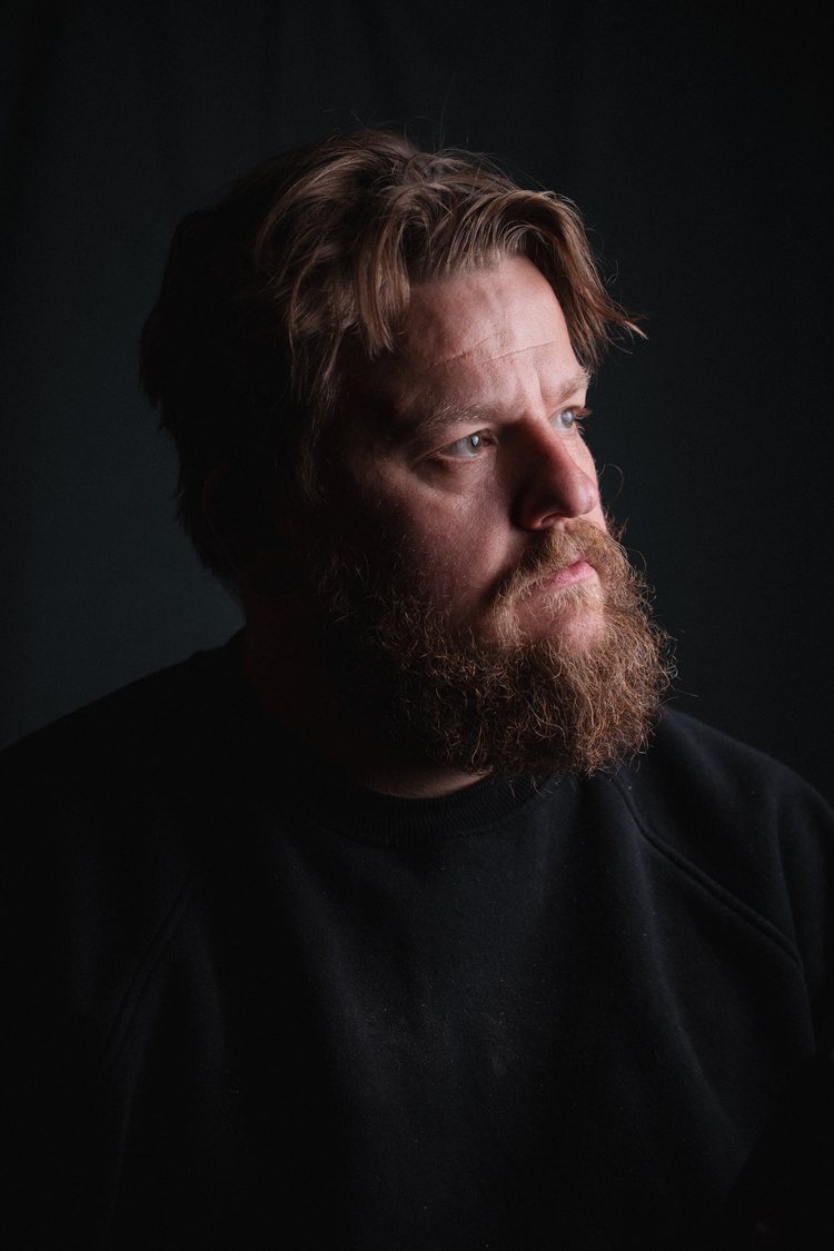 A bearded man in a dark jumper looking to the side in soft light.